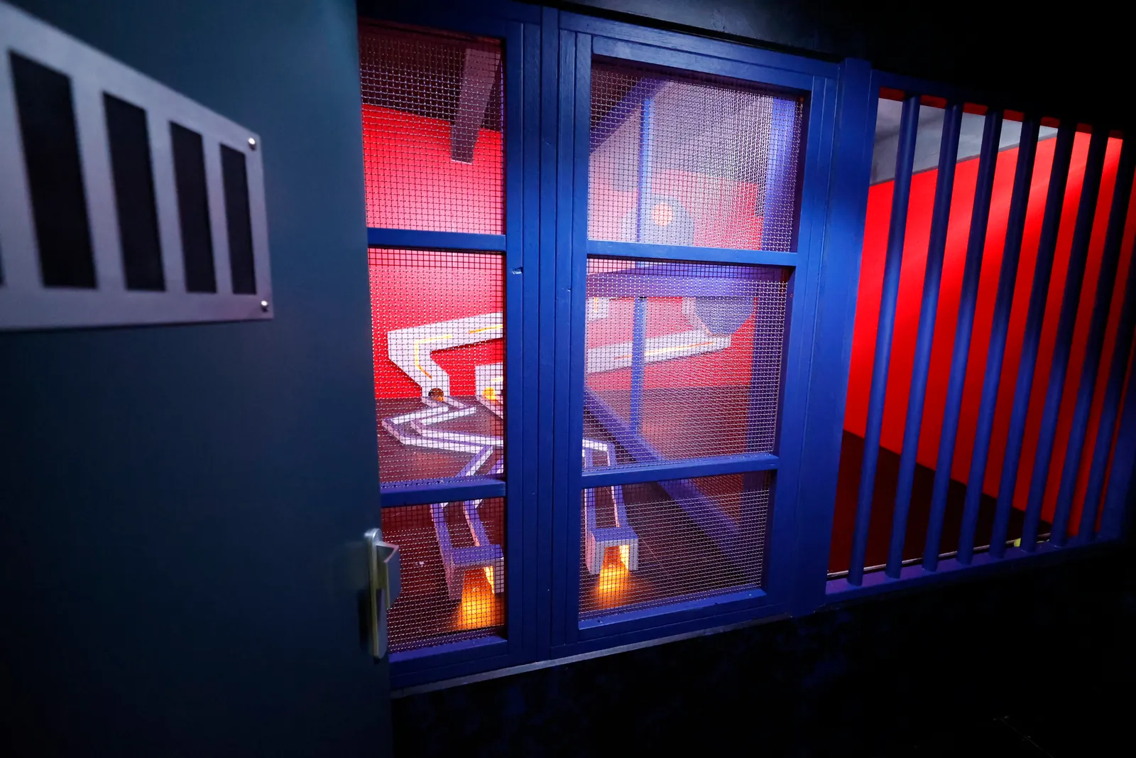 Une fenêtre éclairée en bleu, vue derrière des barreaux rouges